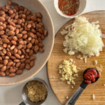 Ingredients laid out for Mexican pinto beans: canned pinto beans, a small white bowl of mixed seasonings, diced white onion, minced garlic, tomato puree, and brown sugar.  Chipotle paste, vegetable stock, and apple cider vinegar are not pictured.