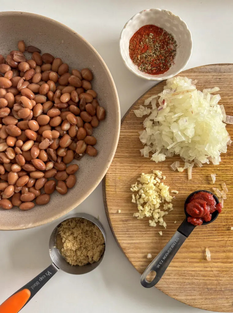 Ingredients laid out for Mexican pinto beans: canned pinto beans, a small white bowl of mixed seasonings, diced white onion, minced garlic, tomato puree, and brown sugar.  Chipotle paste, vegetable stock, and apple cider vinegar are not pictured.