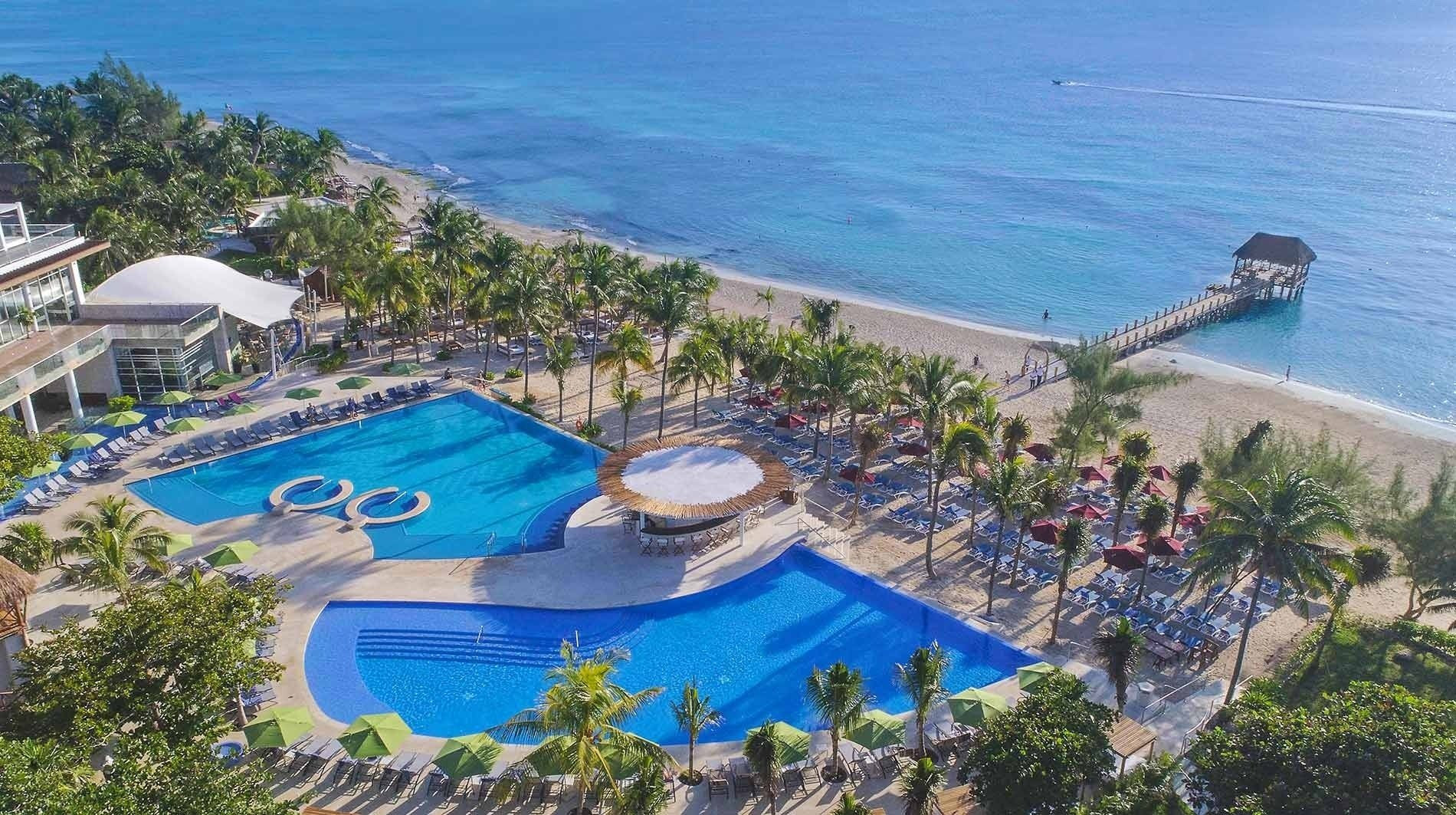 alt= Aerial view of a Cancun beach resort with numerous pools and beach umbrellas.