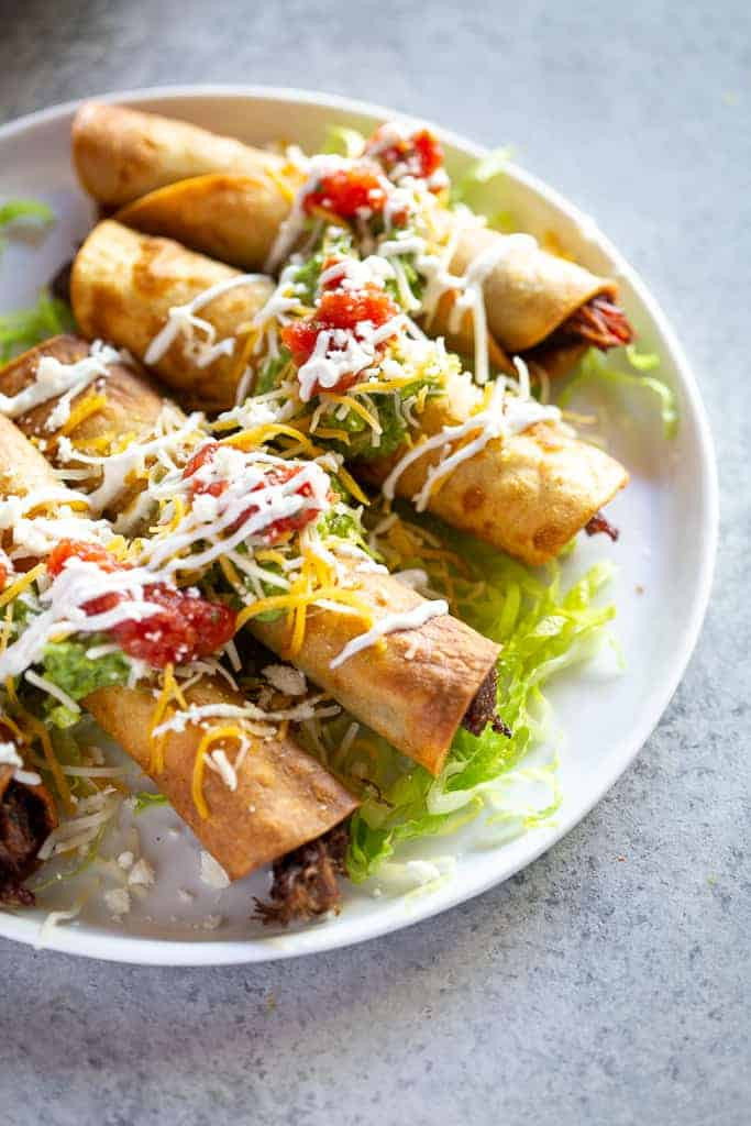 A close-up shot of perfectly topped Mexican taquitos on a plate, featuring guacamole, cheese, sour cream, and salsa, highlighting the delicious and customizable nature of the dish.