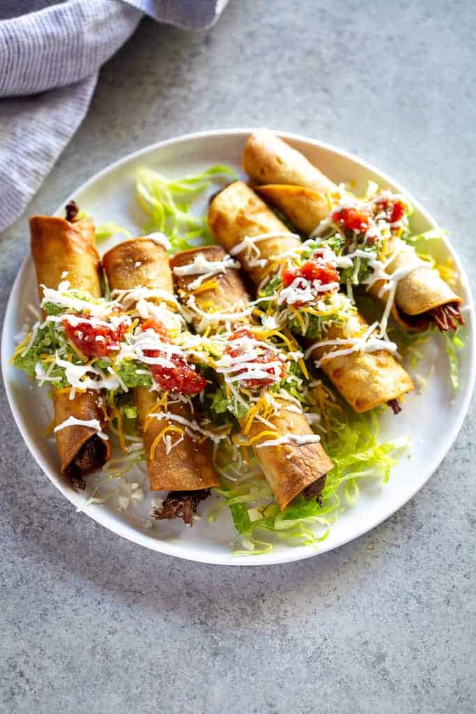 Five beef taquitos beautifully arranged on a plate, topped with guacamole, sour cream, and salsa, showcasing a delicious and authentic Mexican meal.