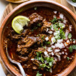 A steaming bowl of beef birria garnished with fresh cilantro, diced onions, and a bright lime wedge, showcasing its rich, red broth and tender meat.