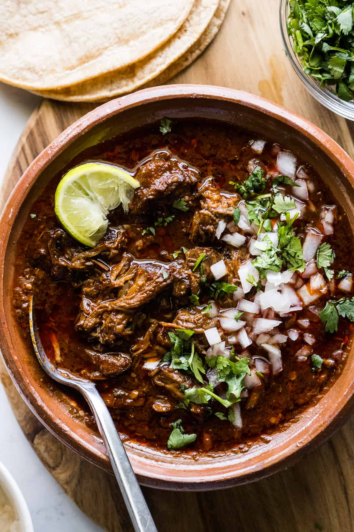 A steaming bowl of beef birria garnished with fresh cilantro, diced onions, and a bright lime wedge, showcasing its rich, red broth and tender meat.