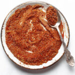 Close up of homemade mexican chicken seasoning on a white plate with a silver spoon, showcasing the texture and color of the spice blend.