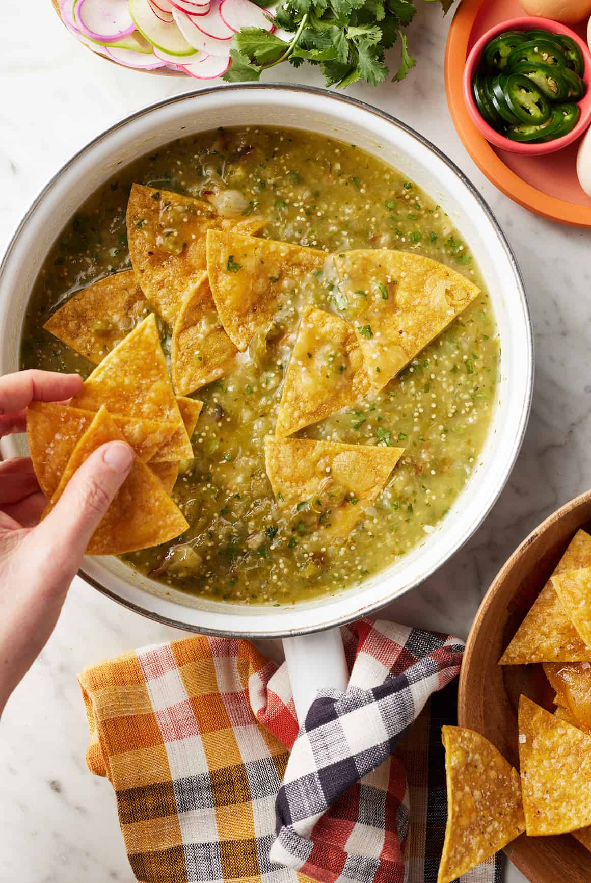 Hand folding crispy tortilla chips into a skillet of salsa verde.