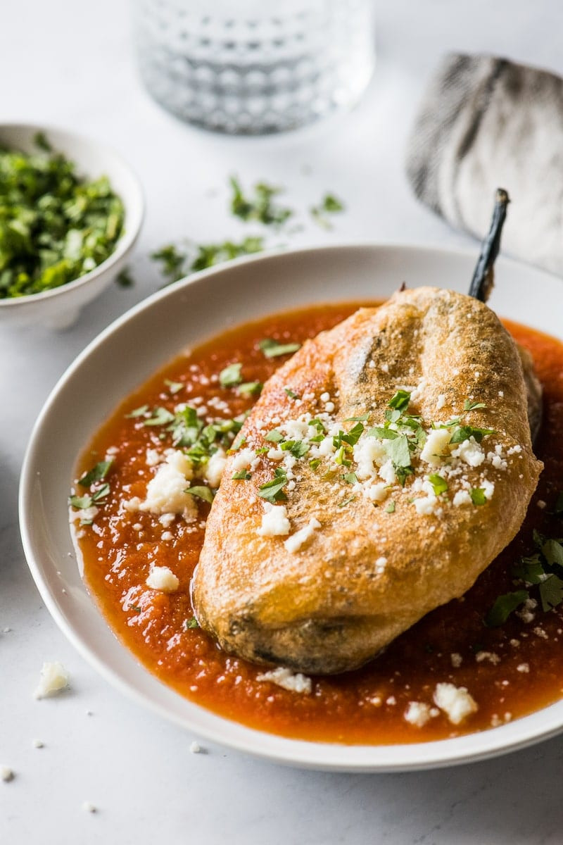 Close-up of a golden brown Mexican chile relleno topped with red salsa on a white plate.