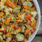 Close-up shot of Doritos Taco Salad in a clear bowl, showcasing layers of lettuce, seasoned ground beef, tomatoes, cheese, Doritos, and dressing.