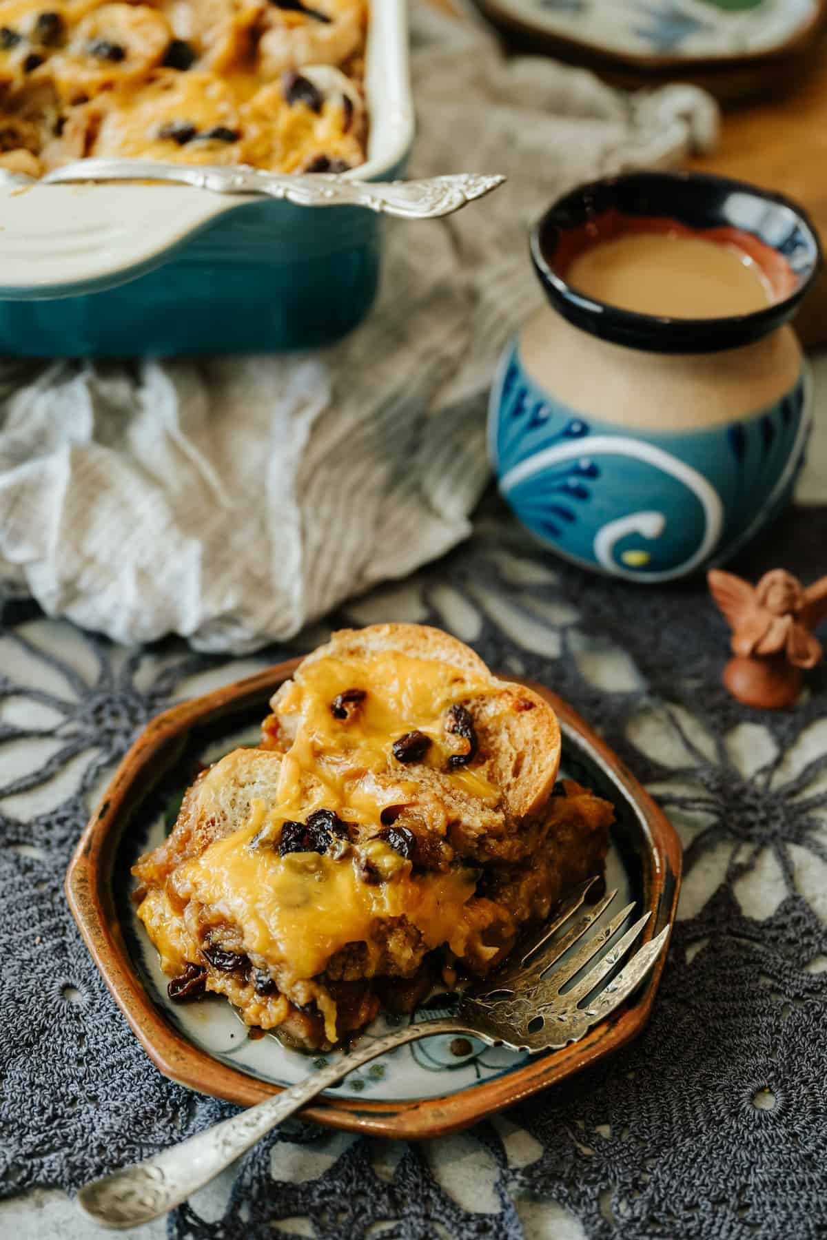 serving of capirotada on a small dessert plate next to a small statue of an angel, a pot of cafe de olla, and the remaining Mexican bread pudding in the casserole dish in the background.