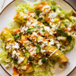 Close-up of golden brown flautas generously topped with crema, hot sauce, salsa verde, fresh lettuce, vibrant cilantro, and crumbled cotija cheese, served on a white plate.