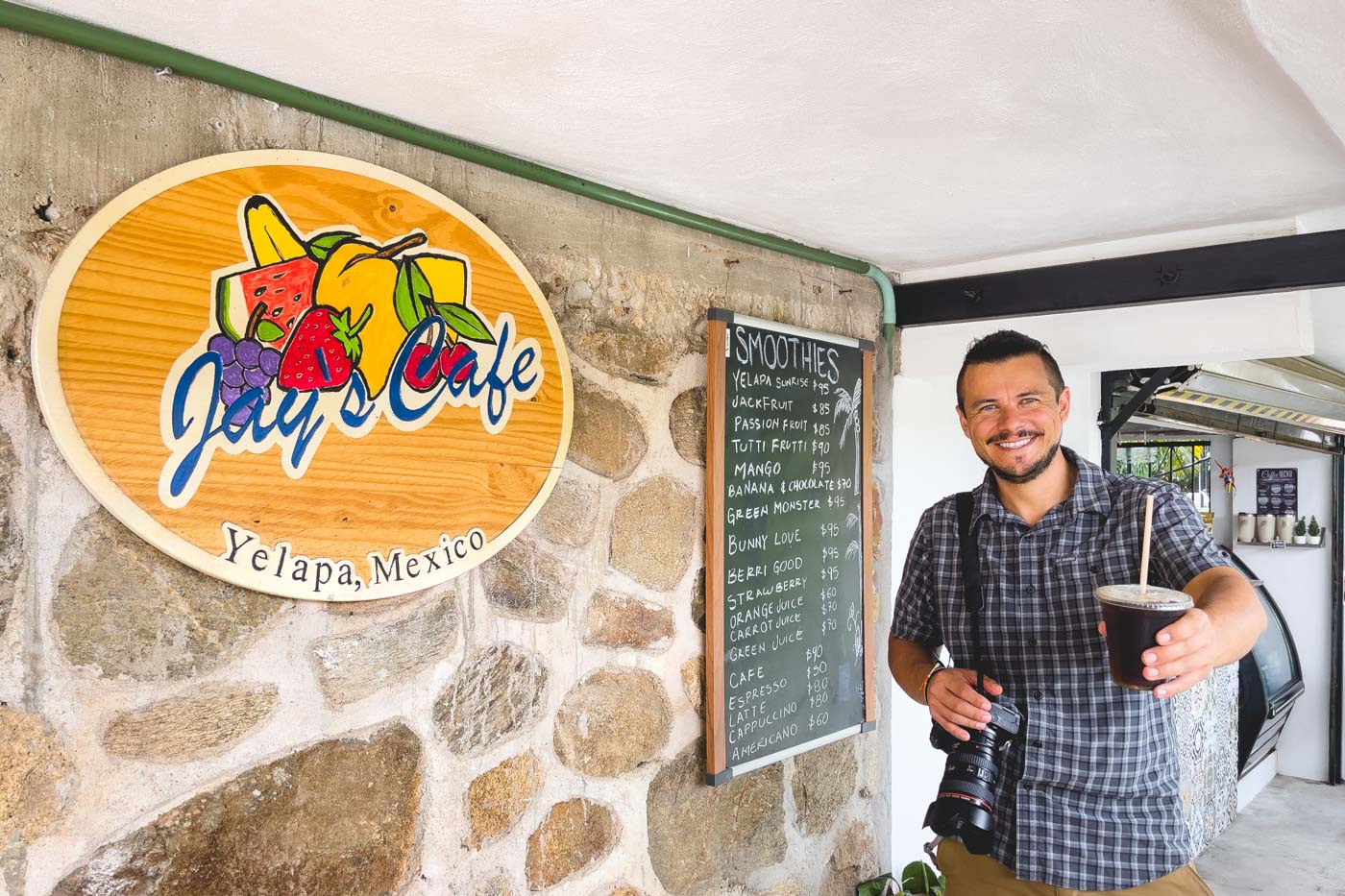 A cheerful man with a camera over his shoulder holding a coffee cup outside Jay's Cafe.