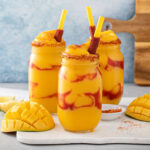 A close-up of a vibrant Mangonada drink, showcasing layers of mango, chamoy, and Tajin seasoning, topped with fresh mango chunks and a tamarind straw, perfect for a hot day.