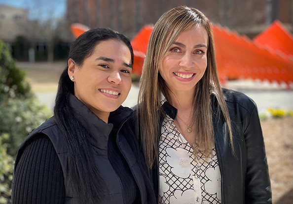 Two female graduate students smiling, standing outside in front of a blurry orange sculpture in the background