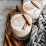 Two glasses of creamy Mexican Horchata filled with ice and garnished with cinnamon sticks and a sprinkle of ground cinnamon.