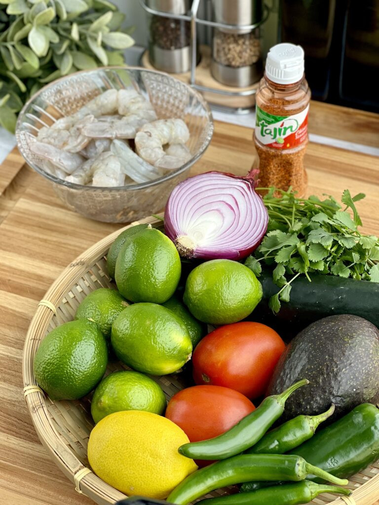 Ingredients for Mexican Shrimp Ceviche laid out on a table, including shrimp, limes, lemons, tomatoes, red onion, cucumber, cilantro, jalapeños, serrano peppers, avocado, and Tajin seasoning.