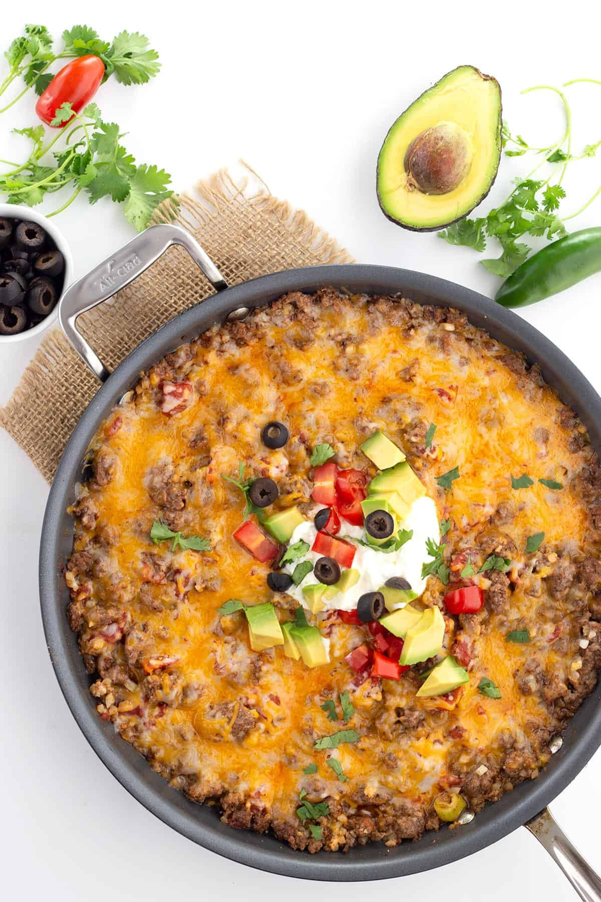 A top-down view of Mexican cauliflower rice in a skillet, garnished with avocado, cilantro, jalapeno, and olives.