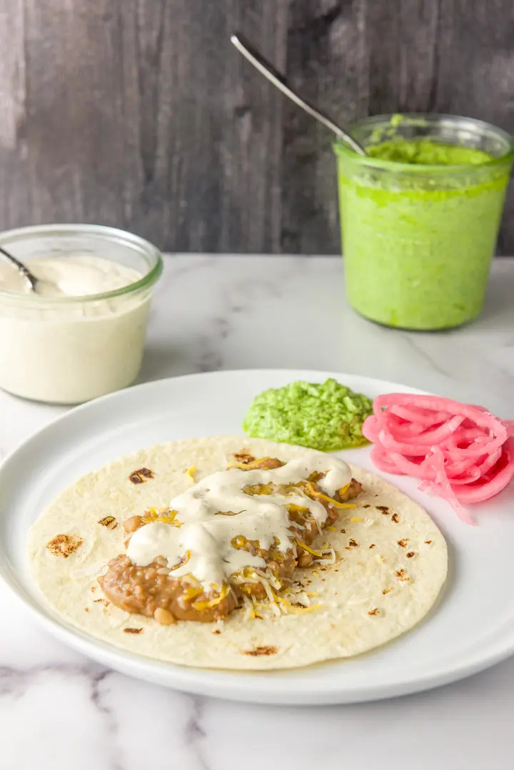 Crema on some beans and cheese in a flour tortilla with pesto, onions and a jar of crema in the background