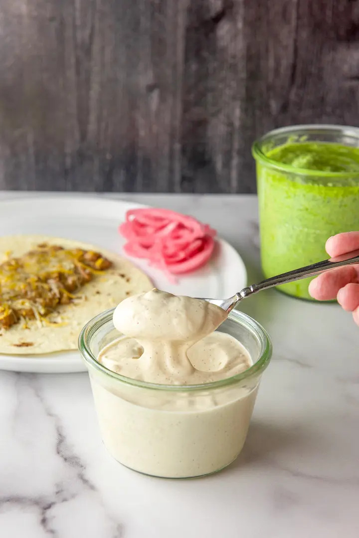 Spoonful of crema over the jar. There is a tortilla covered with beans, onions and pesto in the background