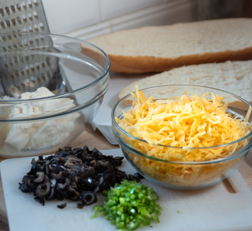 Ingredients for Mexican Garlic Cheese Bread including bread, cheese, mayonnaise, butter, olives, jalapenos, and spices