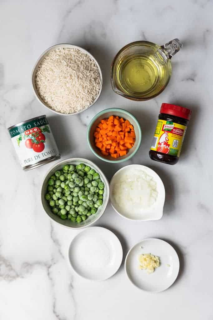 Ingredients for authentic Mexican rice laid out on a wooden surface: rice, tomato sauce, onion, garlic, bouillon, carrots, peas, and serrano peppers.