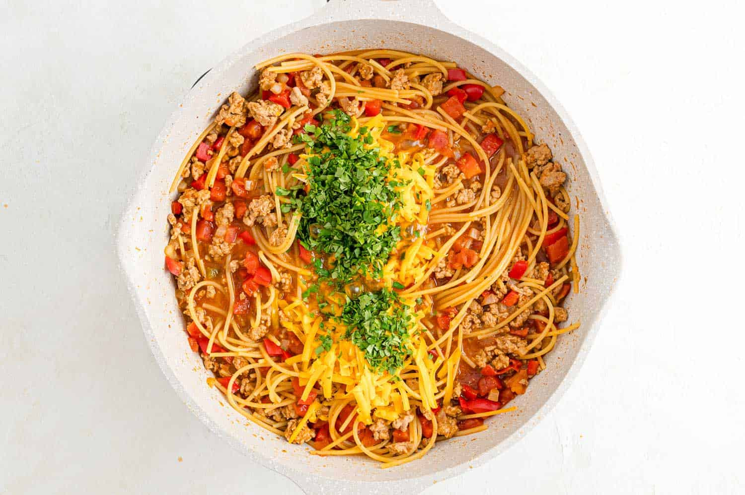 Image of cheese and cilantro being stirred into the cooked Mexican Spaghetti in the skillet, melting into the dish.