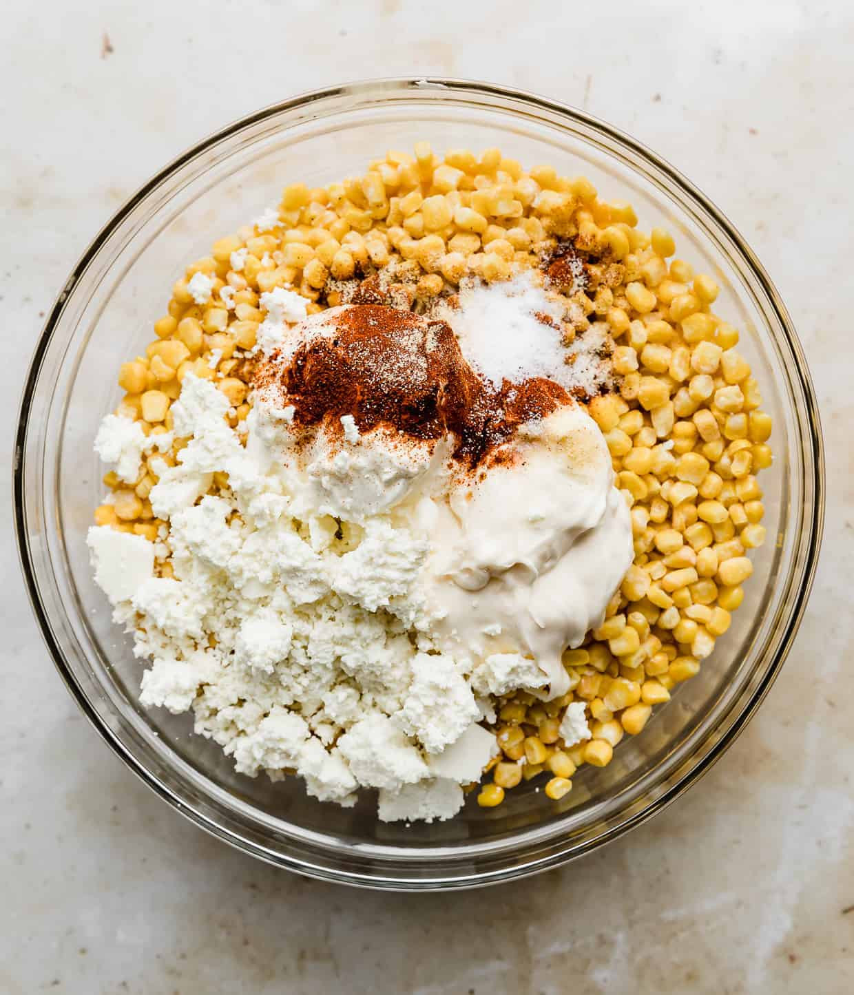 A glass bowl filled with corn kernels, chili powder, mayonnaise, sour cream, and crumbled Mexican cheese, ready to be mixed.