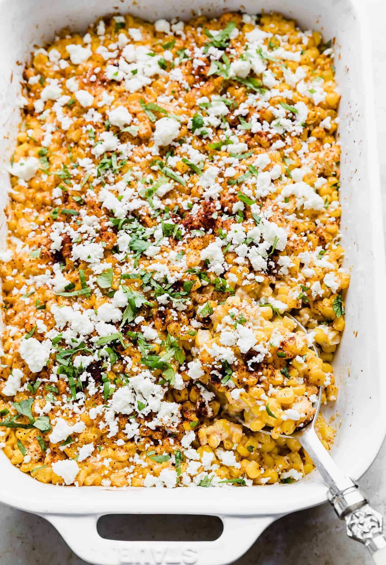 A spoon digging into a warm Mexican Street Corn Casserole, showcasing the creamy texture and visible corn kernels, garnished with cilantro and chili powder.