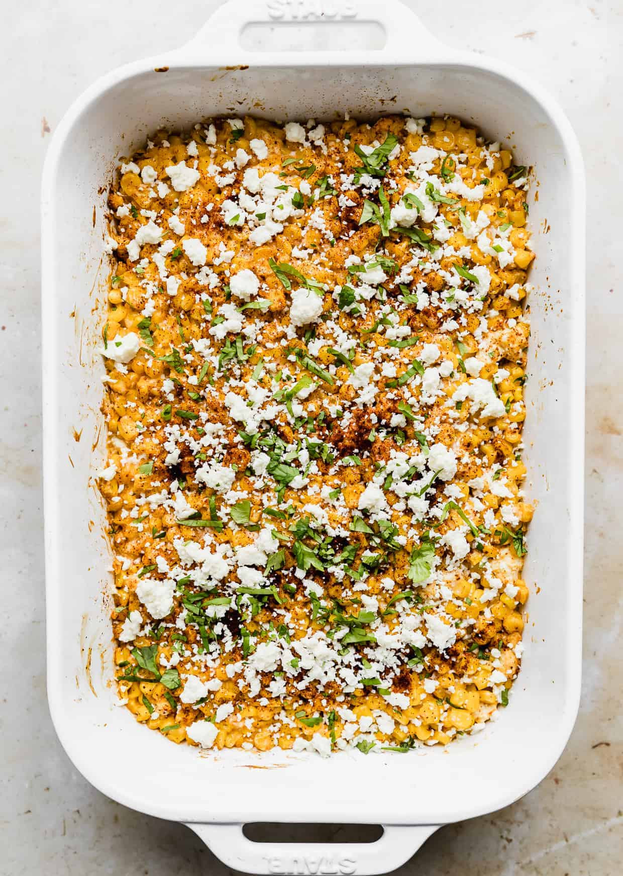 A freshly baked Mexican Street Corn Casserole in a white dish, garnished with fresh cilantro, crumbled white cheese, and a dusting of chili powder, ready to be served.