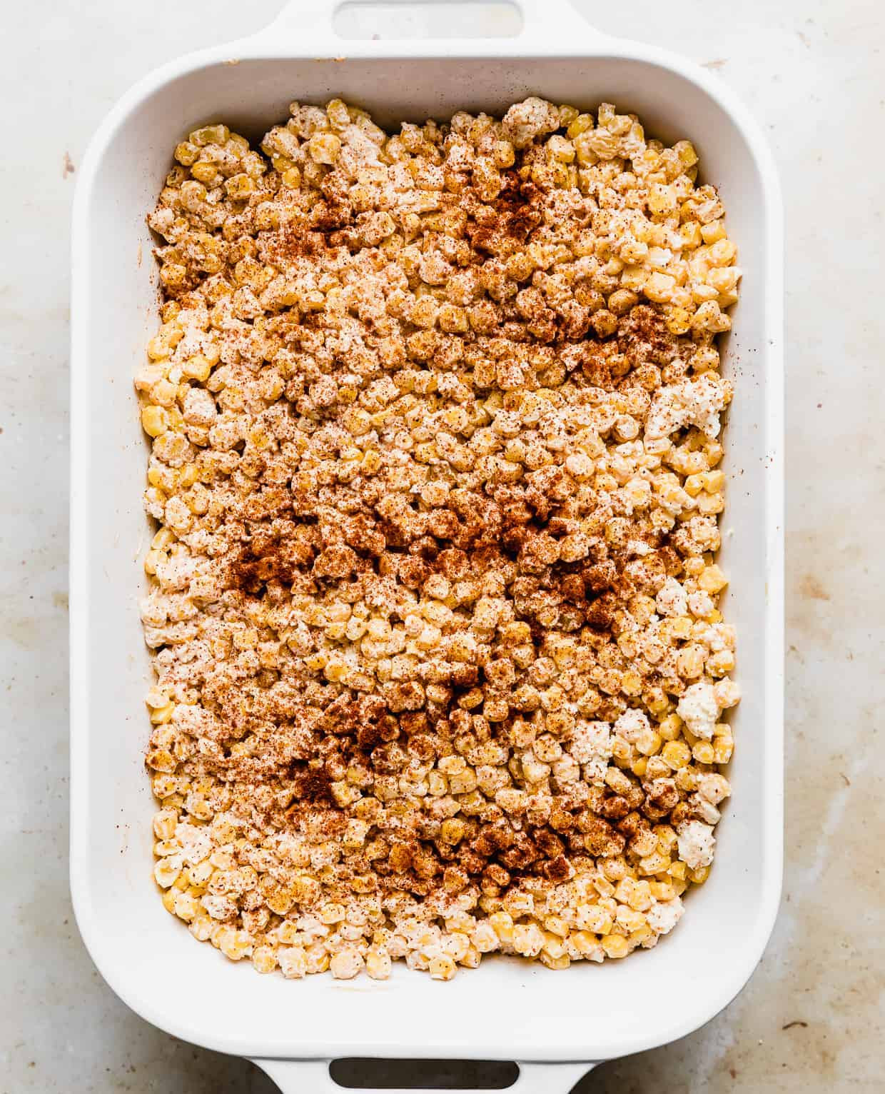 Mexican Street Corn Casserole in a white rectangular baking dish, sprinkled with chili powder before baking, showcasing its vibrant colors.