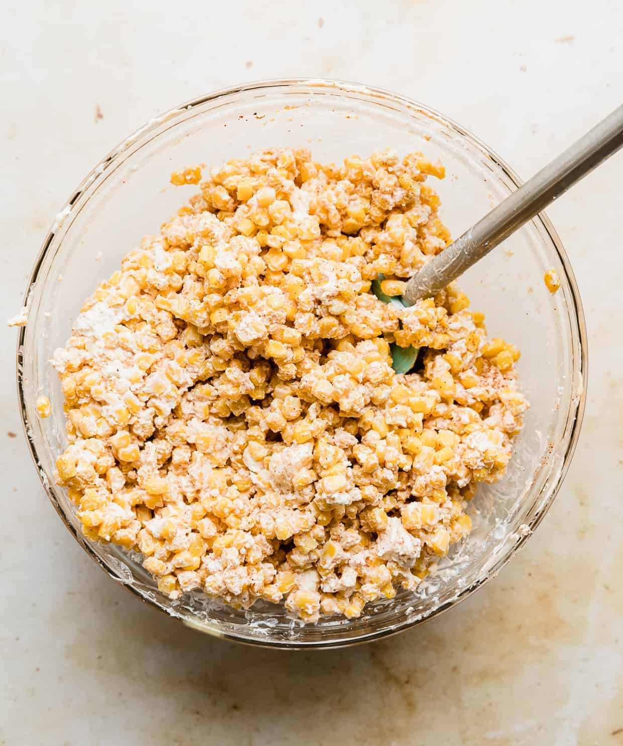 A close-up of corn kernels coated in mayonnaise and sour cream in a large glass bowl, showing the creamy texture of the mixture.