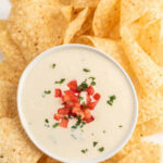 A large white platter filled with tortilla chips with a small bowl of the Mexican white cheese dip in the middle topped with diced tomatoes and fresh cilantro