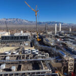 Aerial view of Intel's Fab 9 industrial complex in Rio Rancho, New Mexico, showcasing buildings, pipes, and cranes against a clear sky and mountain backdrop.