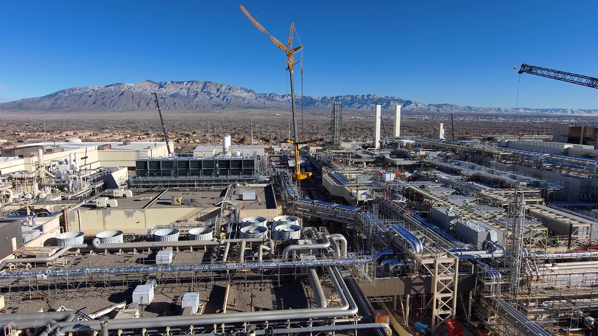 Aerial view of Intel's Fab 9 industrial complex in Rio Rancho, New Mexico, showcasing buildings, pipes, and cranes against a clear sky and mountain backdrop.