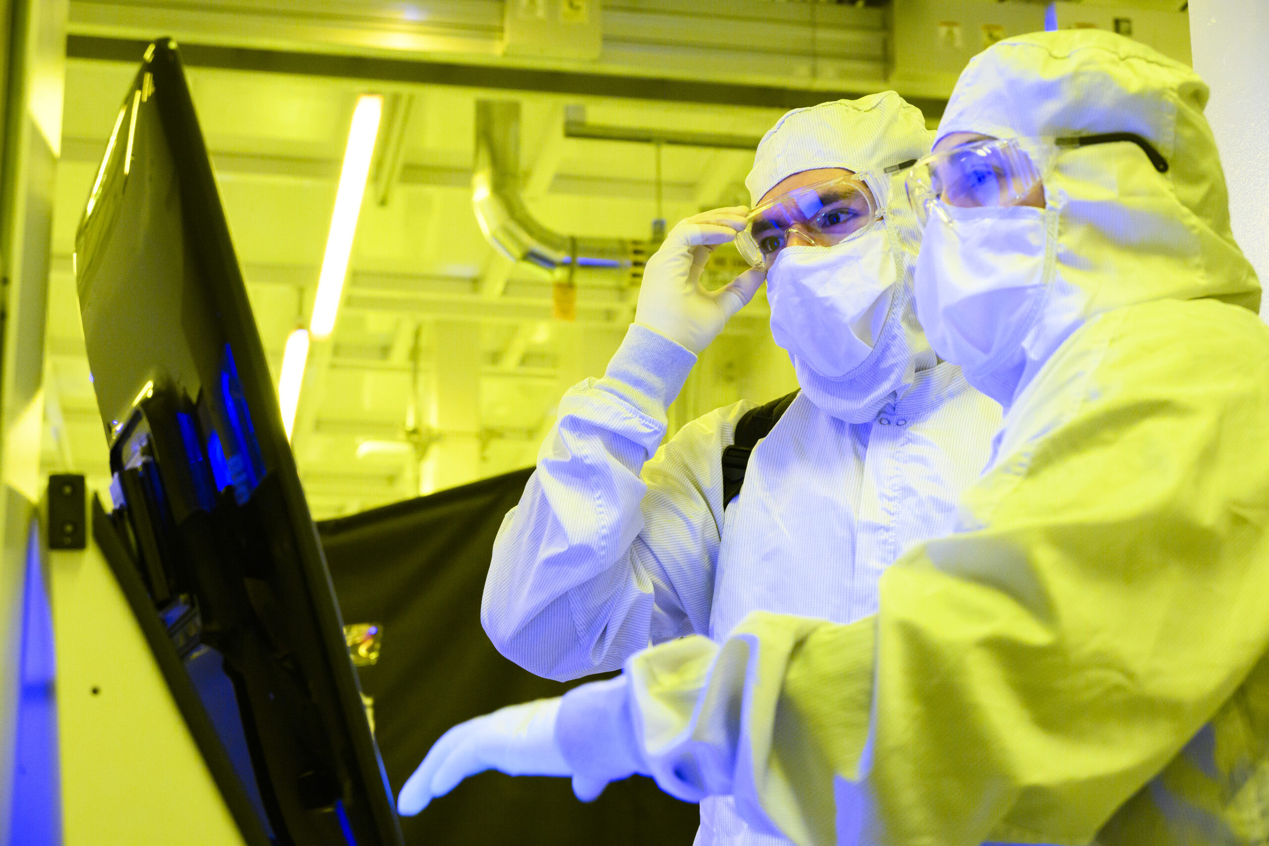 Manufacturing technicians in cleanroom attire work with equipment in Intel's Fab 9, Rio Rancho, New Mexico, under yellow lighting, focusing on advanced semiconductor processes.