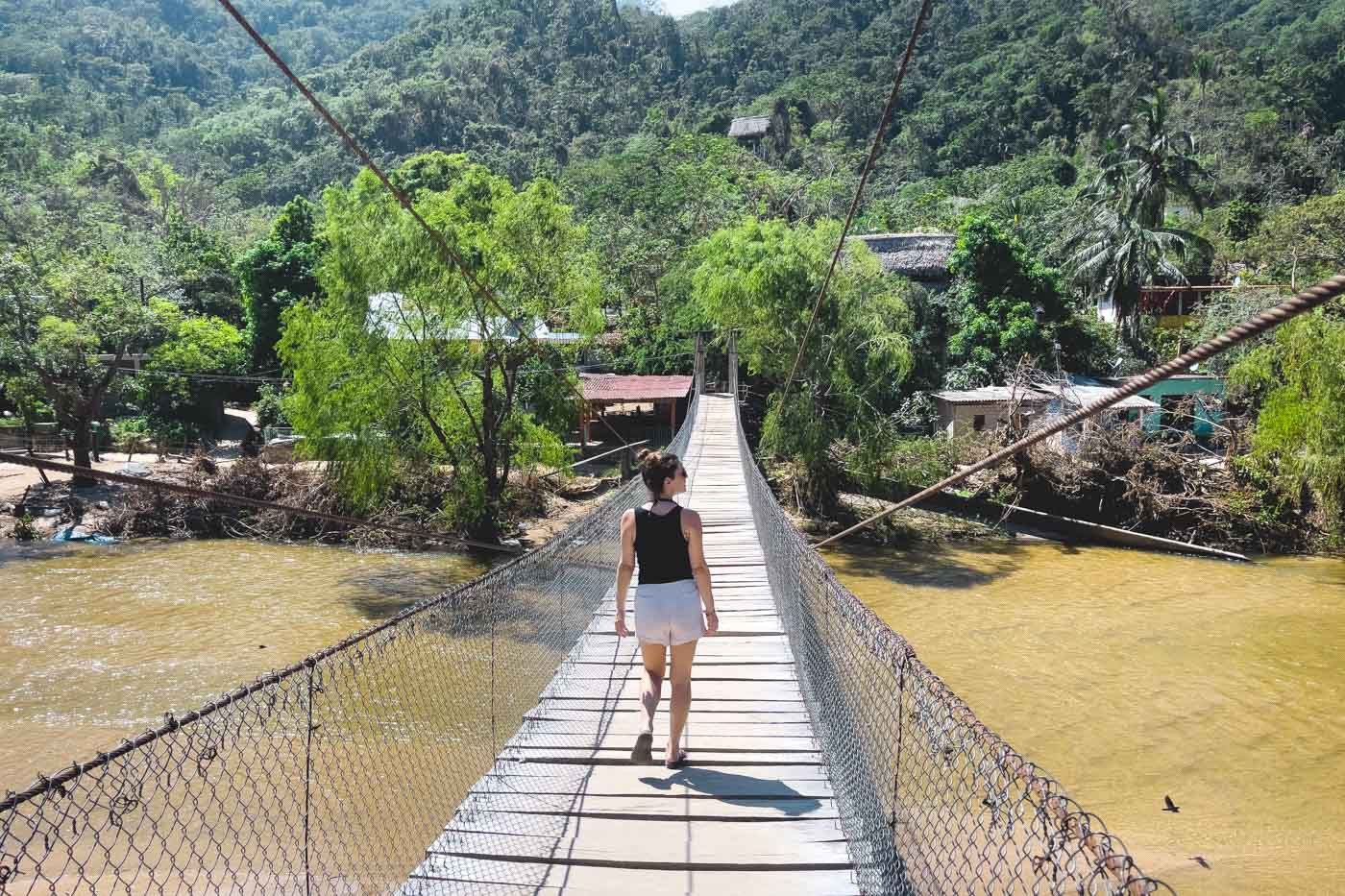 A woman crossing a long wooden suspension bridge over a brown river toward the lush Yelapa forests.