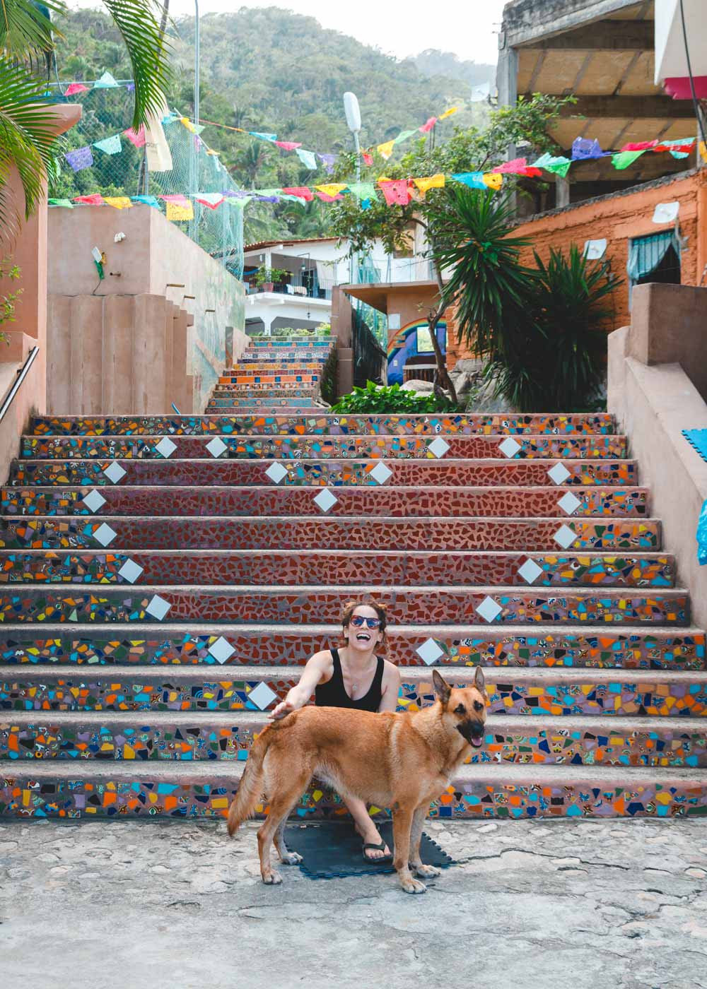 A smiling woman petting a friendly dog on the charming streets of Yelapa, with heart-patterned stairs in the background.
