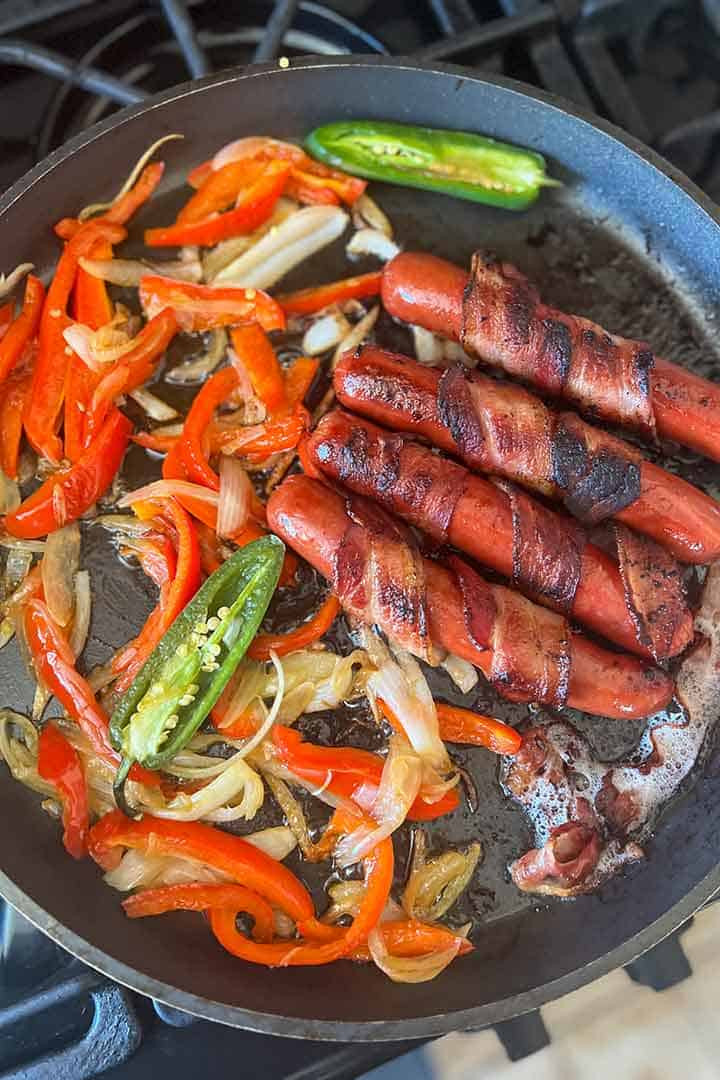 A close-up shot of Mexican hot dogs grilling on a comal, surrounded by sizzling peppers and onions, showcasing the golden-brown bacon and cooked toppings.