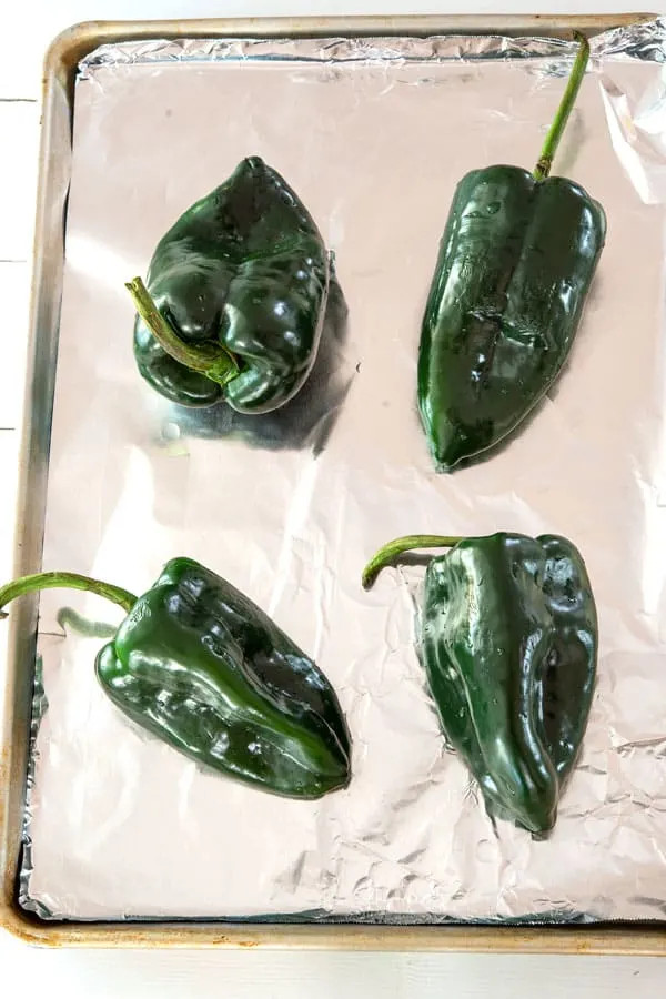 A baking tray with four poblano peppers ready for roasting, demonstrating the initial step of charring the peppers for the sauce