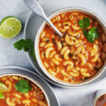 Two bowls of mexican sopa in a vibrant red broth, garnished with lime wedges.