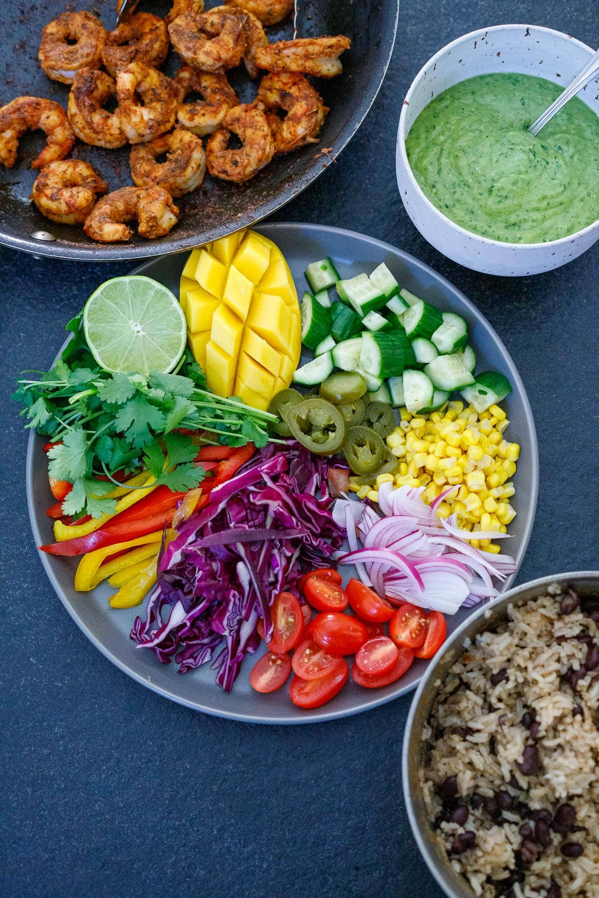 Ingredients for making spicy mexican shrimp and rice bowls, including a skillet of seasoned shrimp, creamy avocado sauce, a plate of chopped vegetables and mango, herbs, and a pot of rice and beans.