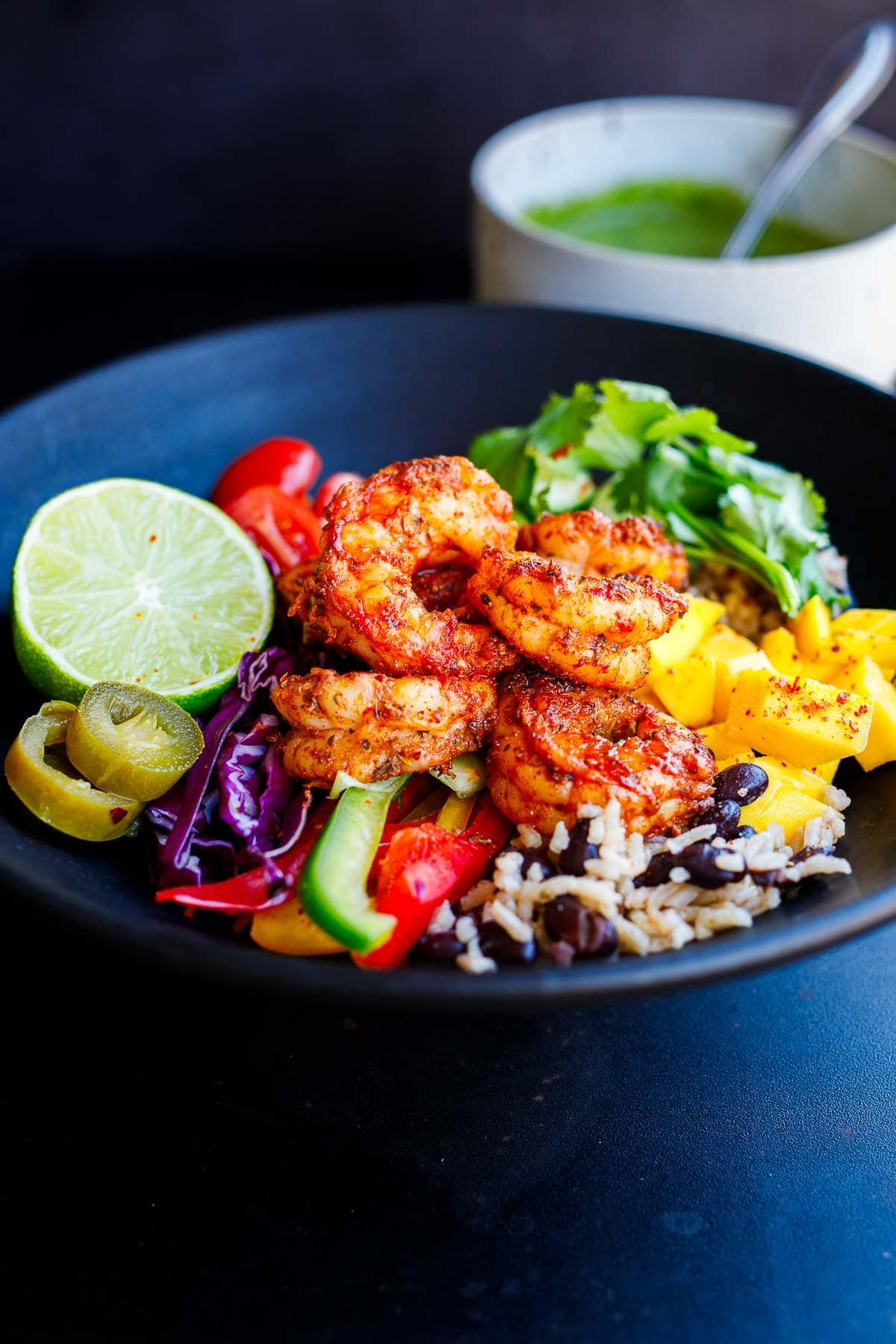Spicy Mexican shrimp served in a rice and bean bowl with a variety of toppings including mango, bell peppers, cabbage, pickled jalapeño, lime, cilantro, and tomatoes.