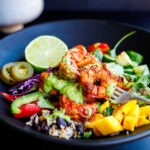 Spicy Mexican shrimp and rice bowl with colorful toppings like mango, bell peppers, cabbage, cucumbers, jalapeno, and lime, all drizzled with creamy avocado sauce.