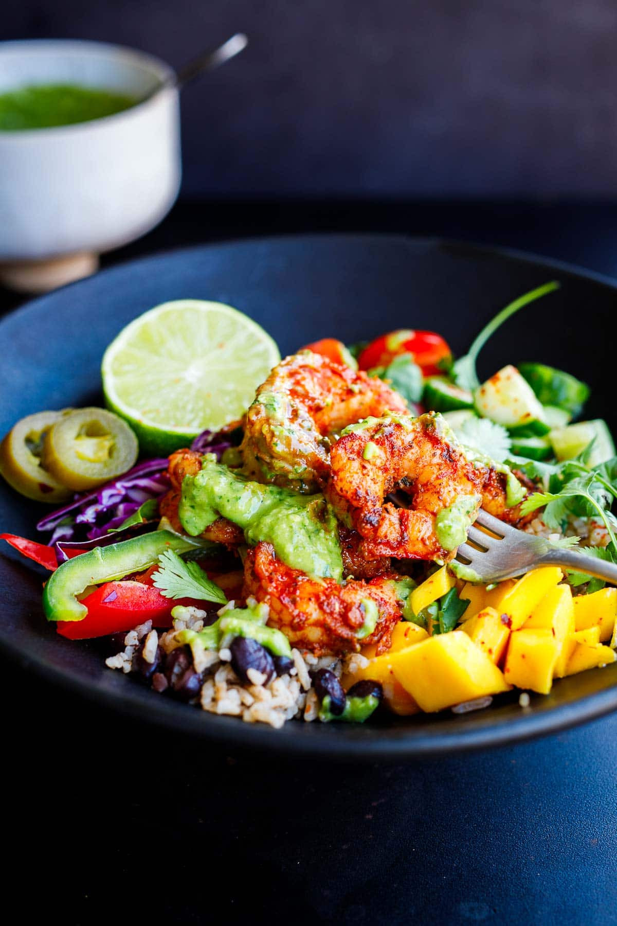 Spicy Mexican shrimp and rice bowl with colorful toppings like mango, bell peppers, cabbage, cucumbers, jalapeno, and lime, all drizzled with creamy avocado sauce.