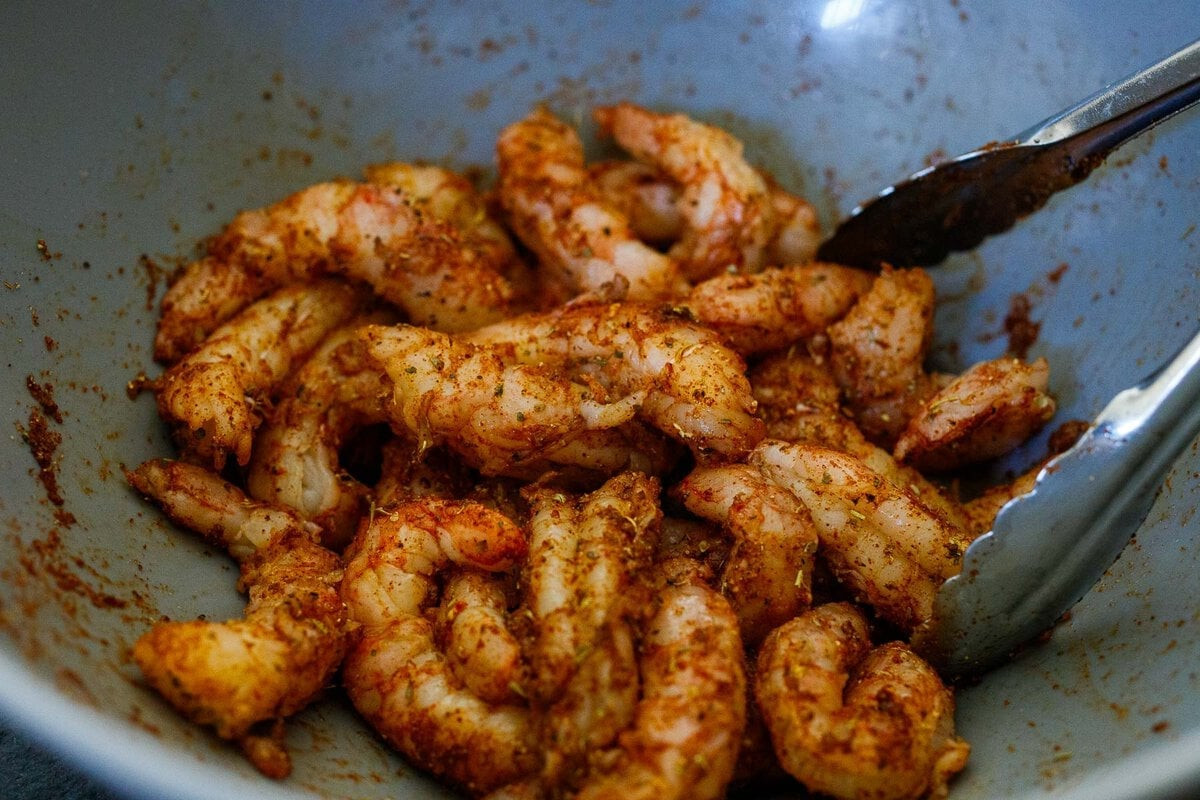 Tongs are used to mix shrimp in a bowl with spicy Mexican seasoning.