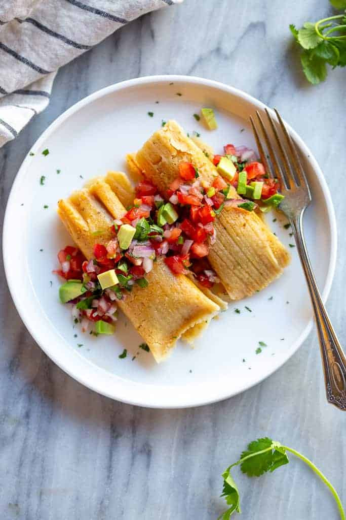 Two tamales on a plate with pico de gallo and chopped avocado.