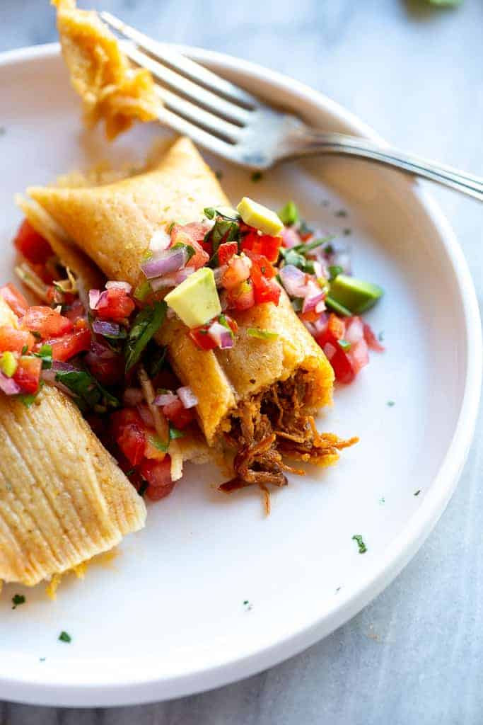 Tamale with pork filling, pico de gallo on top and a fork taken a bite out of it.