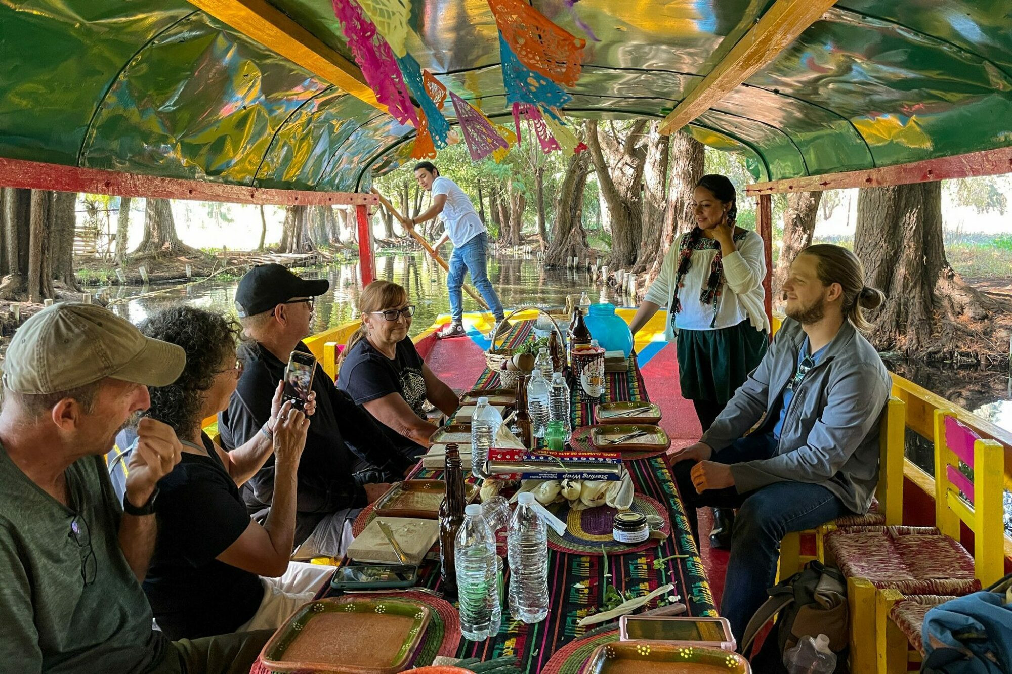 A group enjoying a meal on a trajinera boat in Xochimilco, with food and drinks on a decorated table.