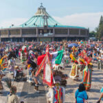 People in traditional costumes, many carrying banners, gather in a square in front of a giant modern church