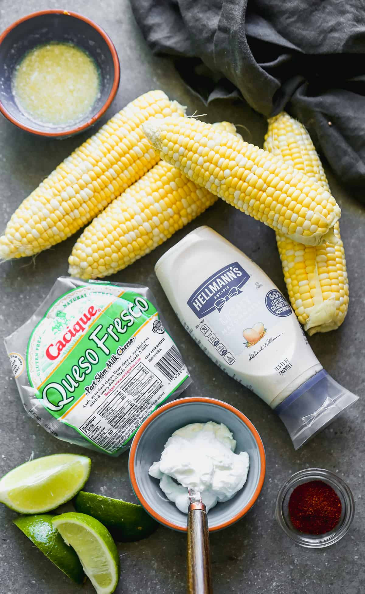 Elote ingredients including corn, mayonnaise, Mexican crema, cotija cheese, chili powder, cilantro, and lime.