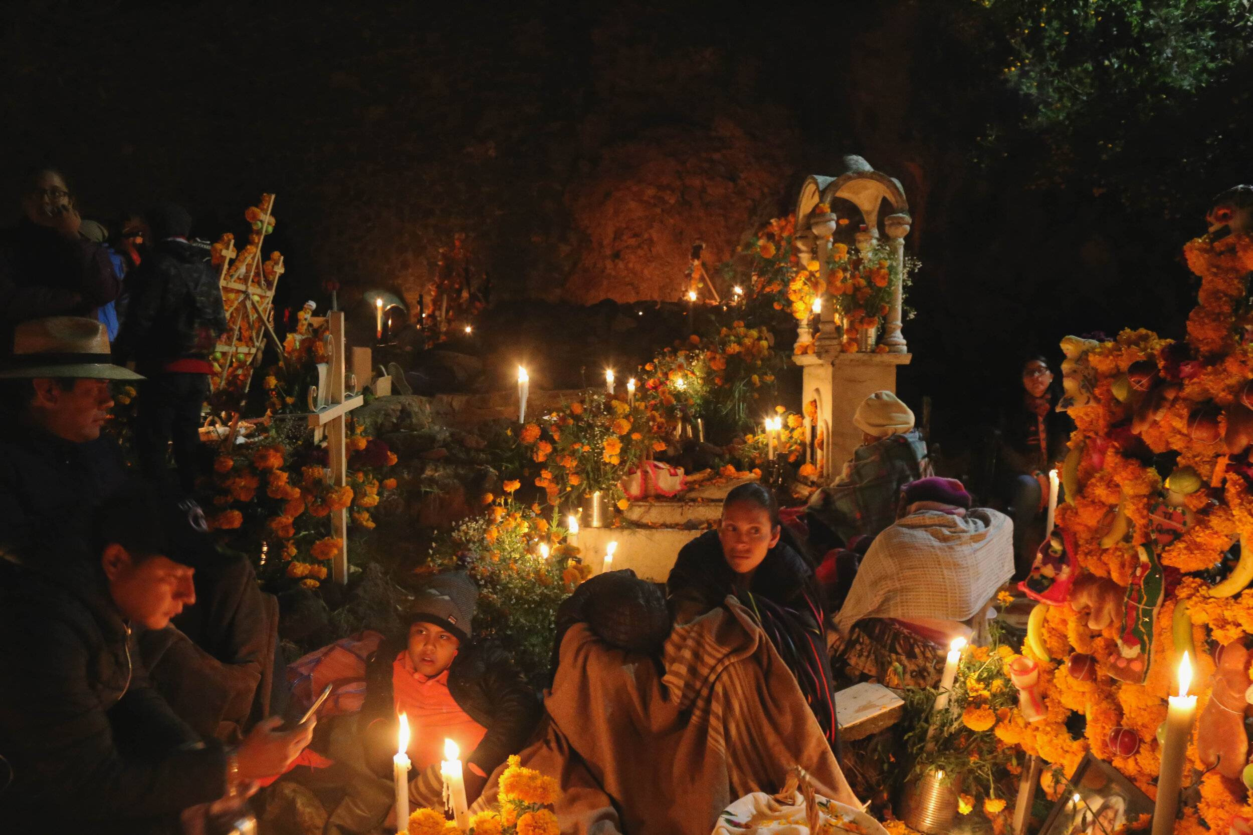 Nighttime scene of people gathered around gravesites, illuminated by numerous candles, reflecting the reverence and cultural significance of honoring deceased loved ones.
