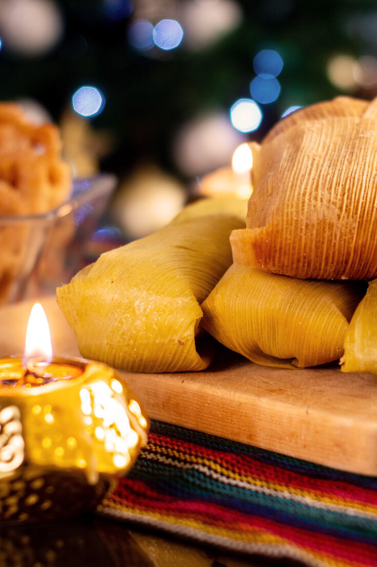 A stack of tamales on a wood board on a table in front of a lit Christmas tree.